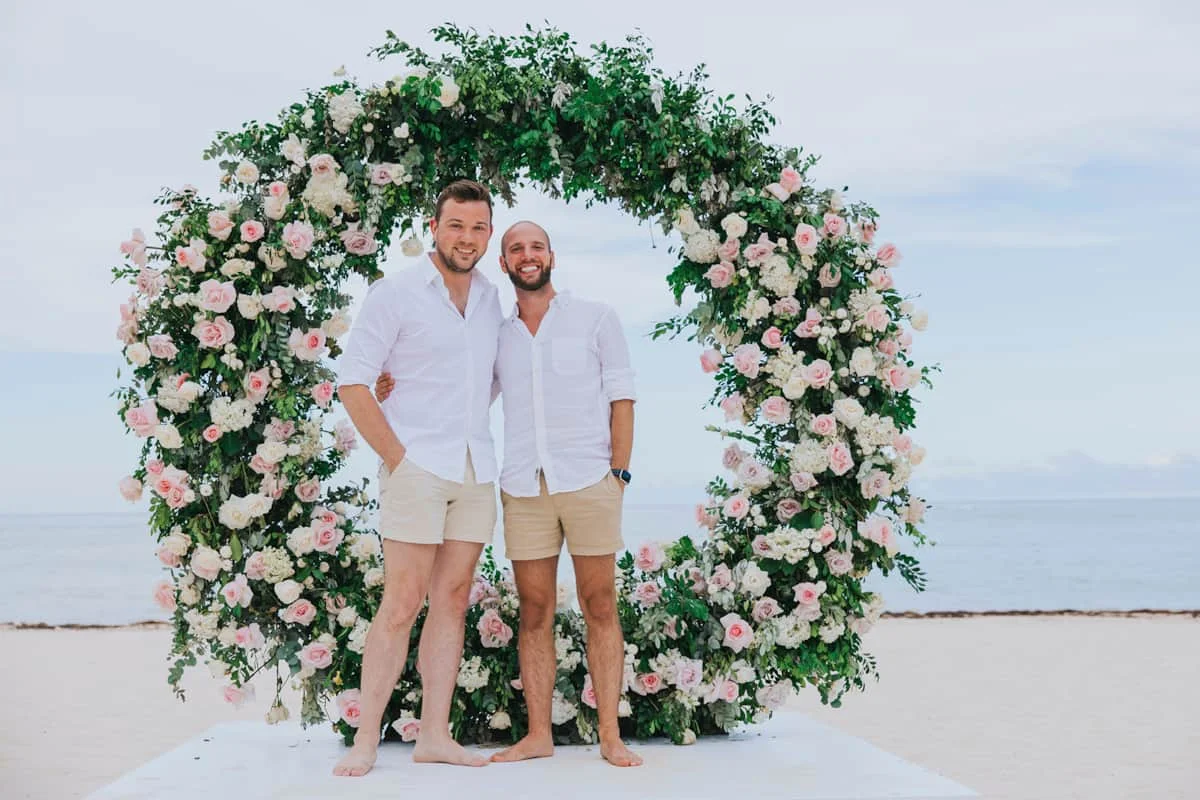 Alex and Hunter, barefoot in white shirts, under a floral arch on their wedding day