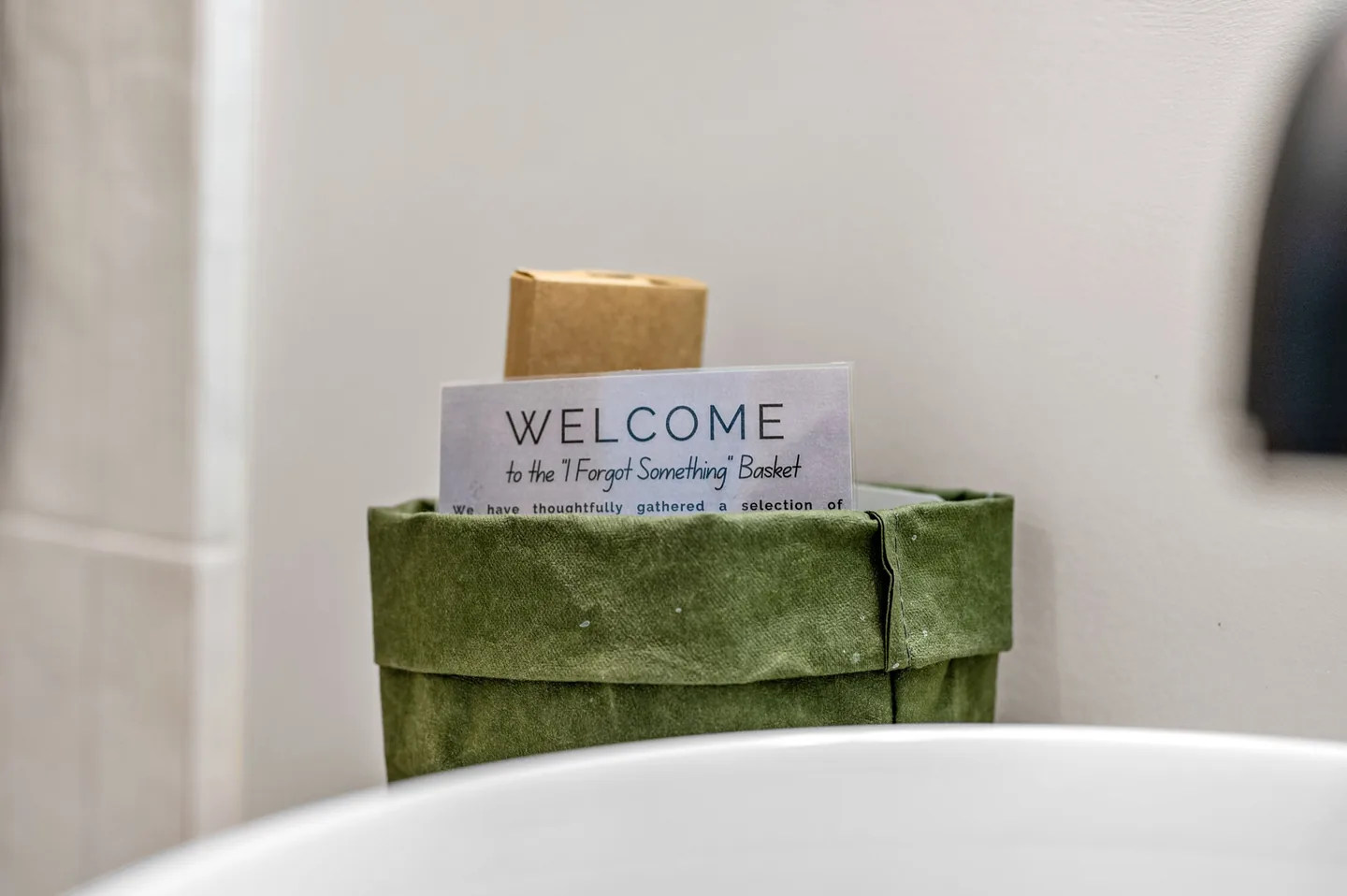 Green paper basket with a welcome card for the “I Forgot Something” basket beside the vessel sink in the guest bathroom