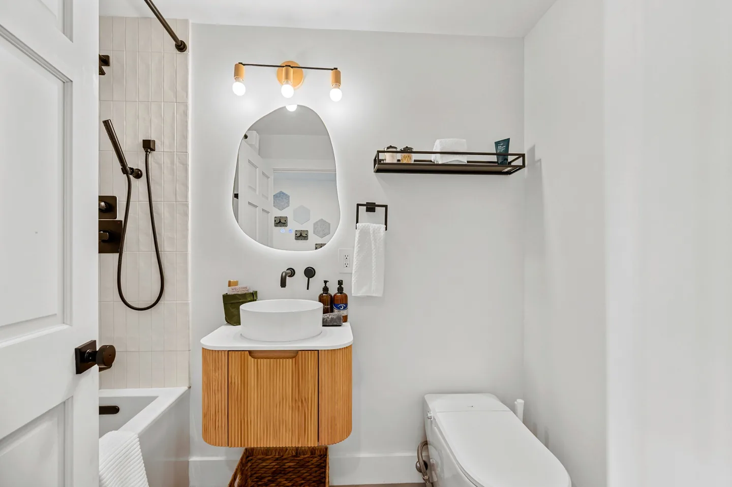 Spa-style guest bathroom with a floating fluted oak vanity, white vessel sink, backlit organic-shaped mirror, black shelf and smart bidet toilet