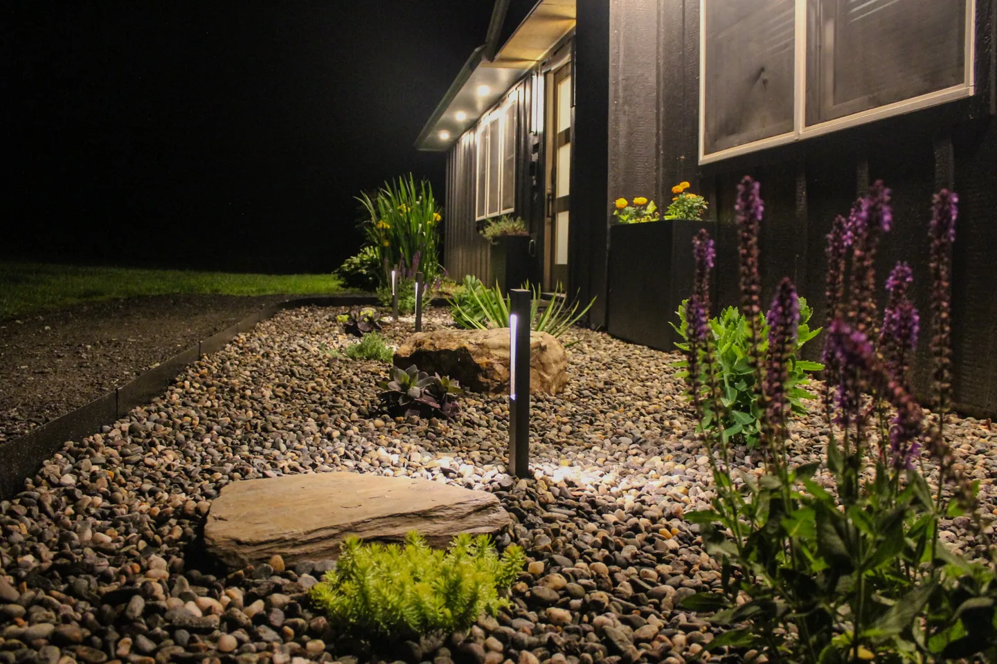 Black board-and-batten Stowe vacation rental entry at night with path lights, a gravel garden, flagstone steps, boulders and purple salvia