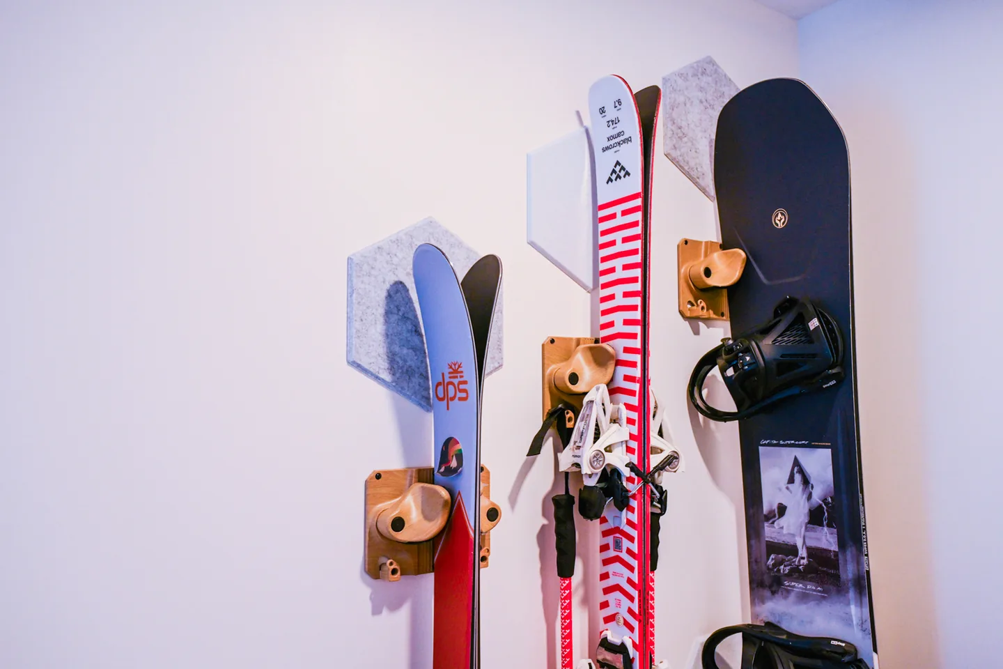 Close-up of wall-mounted wooden ski and snowboard racks with felt hexagon panels holding two pairs of skis and a snowboard